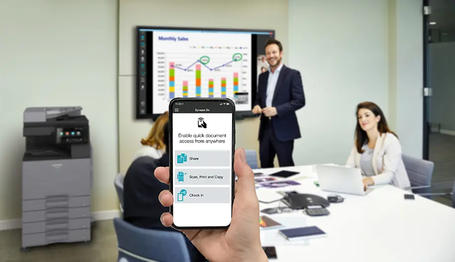 A hand holding a mobile phone displaying the Synappx Go application interface and using the application to select and share content from their mobile phone or cloud storage on a Sharp Display during a meeting in a conference room