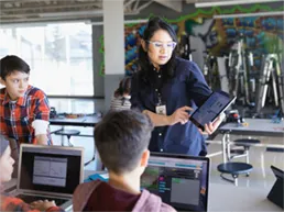 Classroom with teacher pointing at content on a tablet display and 3 students surrounding her looking at laptops