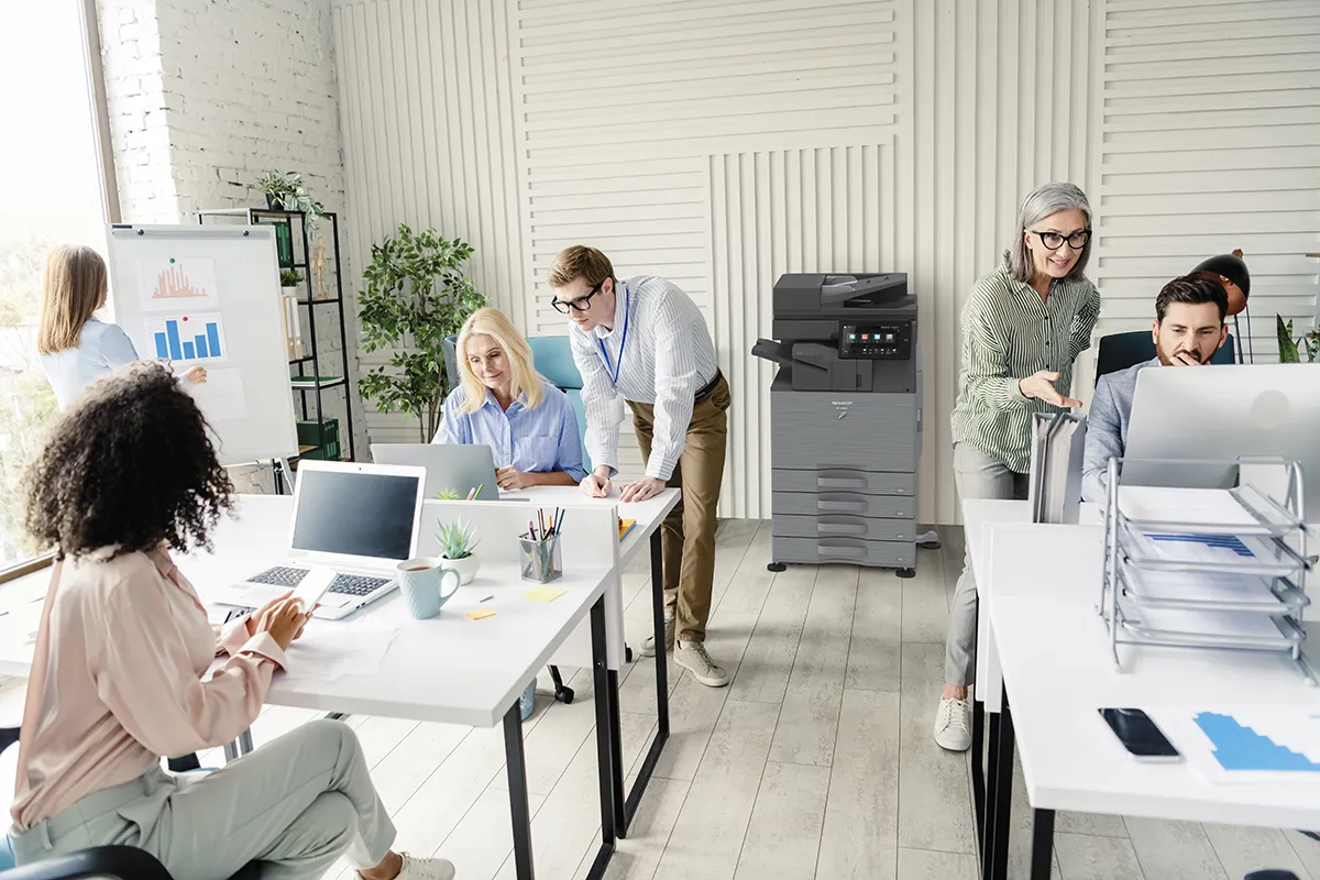 A busy, modern open-plan office scene featuring a group of diverse professionals working and collaborating. In the center, a dark gray Sharp multi-function printer stands against a white-paneled wall. 