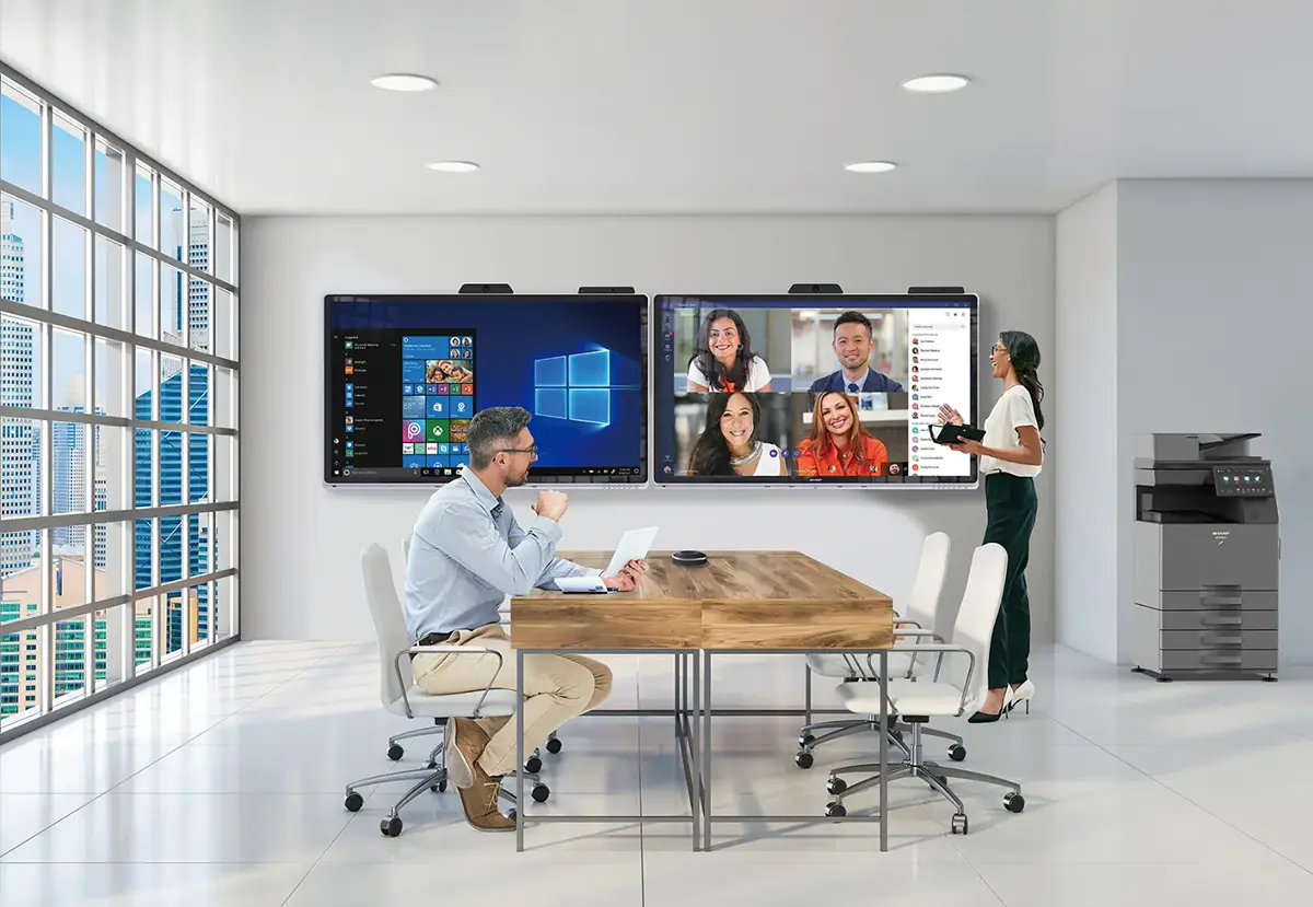 A woman and a man engaging in a video conference call in a meeting room with a Sharp multifunction printer next to the displays.