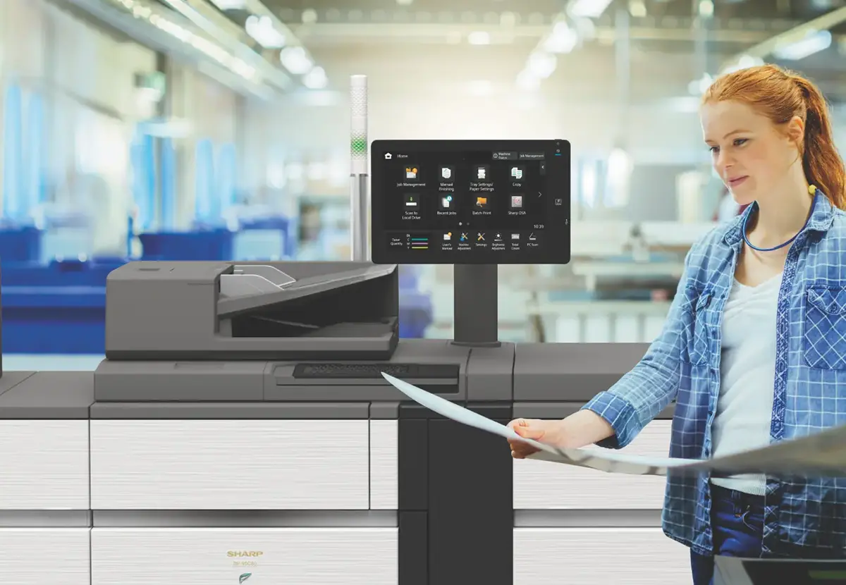 A woman working at a commercial printer stands in front of a Sharp Pro Series Printing System reviews a large printed document
