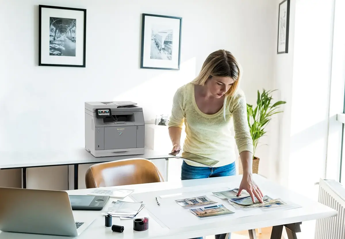 A woman reviewing printed photos on her desk with a compact printer is sitting on the table behind the desk. 
