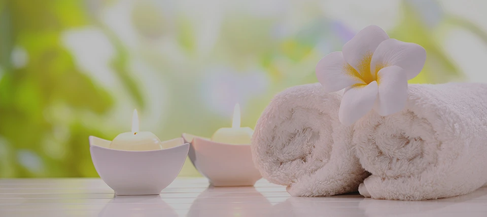 A serene arrangement featuring two rolled white towels, a delicate white flower, and small candles, all set against a softly blurred natural background.