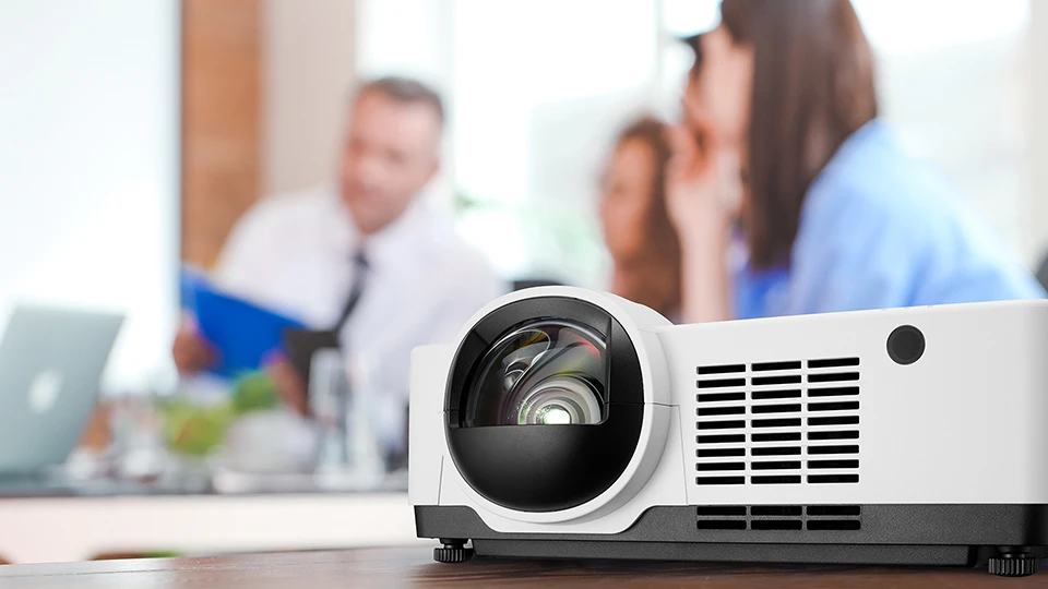 A Sharp laser projector sits on a wooden table in the foreground, while people engaged in discussion are blurred in the background.