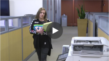 Image of woman walking in office with folder in her arms towards a Sharp multifunction printer