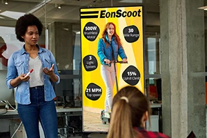A woman with curly hair is giving a presentation about EonScoot, featuring a large screen display with specifications like 500W motor, 30-mile range, and a top speed of 21 MPH.