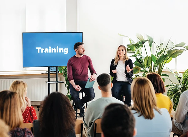 A training session with two speakers in front of an audience, a large screen display behind them displaying the word 'Training'.