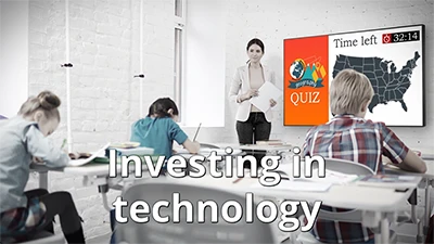 Investing in technology - a teacher is standing in front of a classroom of students taking a quiz with large format display hanging on the wall displaying a map of the United States with the words Geography quiz and a countdown timer on the screen